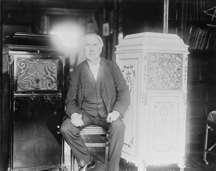 Thomas Alva Edison, full-length portrait, seated, facing front, between two phonograph cabinets, one of which is made of concrete.