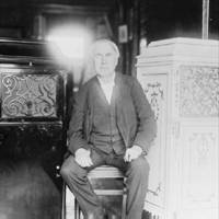 Thomas Alva Edison, full-length portrait, seated, facing front, between two phonograph cabinets, one of which is made of concrete.