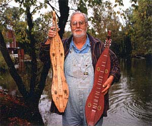 Photo of man with two dulcimers standing outside near water and trees