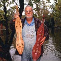 Photo of man with two dulcimers standing outside near water and trees