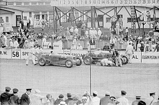 Photo of automobiles and mechanics on a racetrack with spectators watching