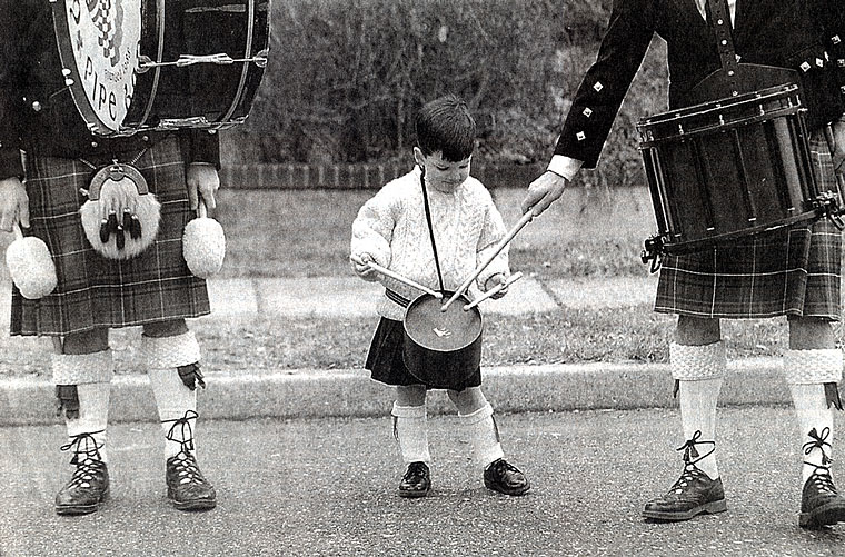 Photo of a young boy playing a drum between two members of the Bergen Pipe Band