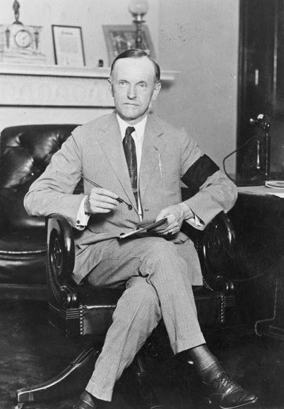 Calvin Coolidge, full-length portrait, seated at desk, facing front, holding pen and paper, wearing black armband in mourning for President Harding, Aug. 4, 1923.