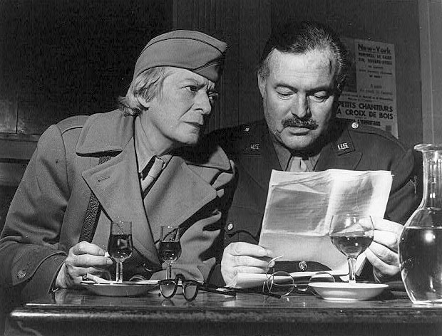 Janet Flanner and Ernest Hemingway, both in uniform, seated reading papers at a table in the Deux Magots cafe in Paris, France.
