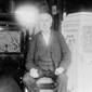 Thomas Alva Edison, full-length portrait, seated, facing front, between two phonograph cabinets, one of which is made of concrete.