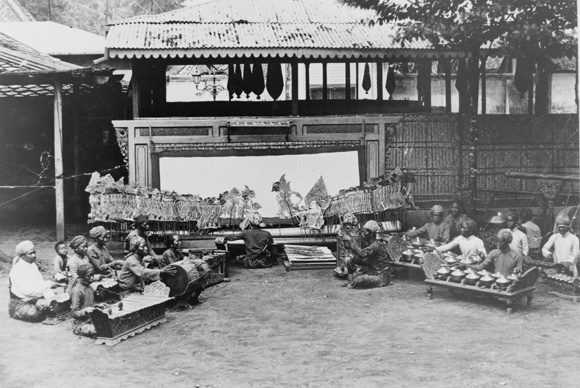 Black and white photo of people sitting on ground behind a stage