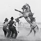 Photo of several people trying to saddle a wild horse while two men on horseback watch from the distance