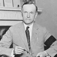 Calvin Coolidge, full-length portrait, seated at desk, facing front, holding pen and paper, wearing black armband in mourning for President Harding, Aug. 4, 1923.