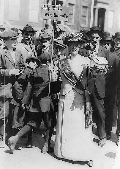 Suffragist, 'Mrs. Suffern,' holding sign; crowd of boys and men behind.